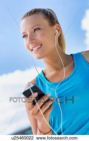 Stock Image - Portrait of young attractive woman standing while using and listening to music on her smartphone in park. Fotosearch