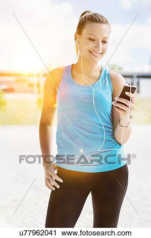 Stock Image - Portrait of young attractive woman standing while using and listening to music on her smartphone in park. Fotosearch