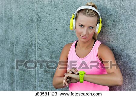 Stock Image - Portrait of young attractive woman leaning on wall wearing sports wear and headphones on. Fotosearch