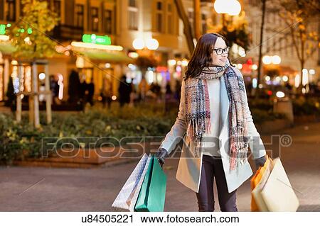 Portrait of young attractive woman walking while holding paper bags on night shopping View Large Photo Image Stock Image - Portrait of young attractive woman walking while holding paper bags on night shopping. Fotosearch
