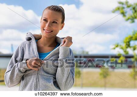 Portrait of young attractive woman smiling while standing and zipping her jacket in park View Large Photo Image Stock Photo - Portrait of young attractive woman smiling while standing and zipping her jacket in park. Fotosearch