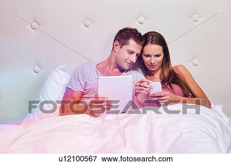 Stock Photo - Portrait of young attractive woman showing smartphone while his boyfriend playing games on his digital tablet in bed. Fotosearch