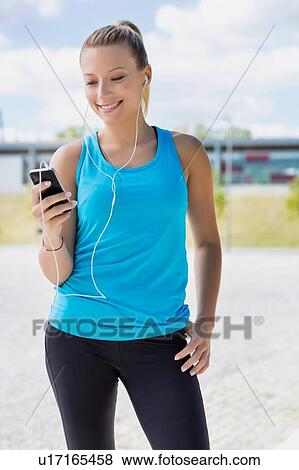 Stock Photo - Portrait of young attractive woman standing while using and listening to music on her smartphone in park. Fotosearch
