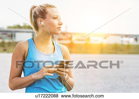 Stock Photo - Portrait of young attractive woman standing while using and listening to music on her smartphone in park. Fotosearch