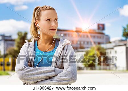 Stock Photo - Portrait of young attractive woman standing with arms crossed while listening to music on her smartphone in park. Fotosearch