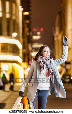 Stock Photo - Portrait of young attractive woman standing while calling for taxi after night shopping. Fotosearch