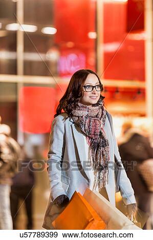 Stock Photo - Portrait of young attractive woman walking while holding paper bags on night shopping. Fotosearch