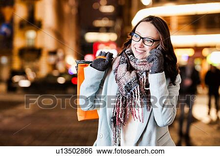 Portrait of young attractive woman smiling and talking on smartphone while holding paper bags at night shopping View Large Photo Image Stock Photograph - Portrait of young attractive woman smiling and talking on smartphone while holding paper bags at night shopping. Fotosearch