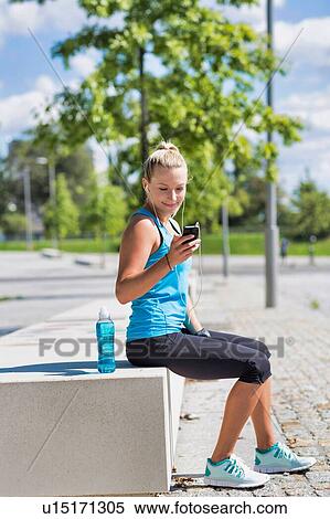 Stock Photography - Portrait of young attractive woman sitting while using and listening to music on her smartphone in park. Fotosearch