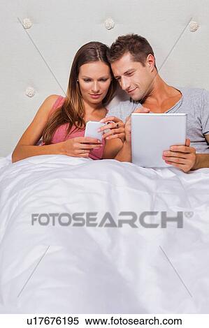 Stock Photography - Portrait of young attractive woman showing smartphone while his boyfriend playing games on his digital tablet in bed. Fotosearch