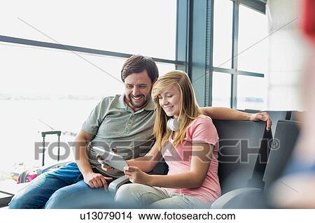 Picture - Portrait of young beautiful teenage girl showing digital tablet to her father while sitting and waiting for their flight in airport. Fotosearch
