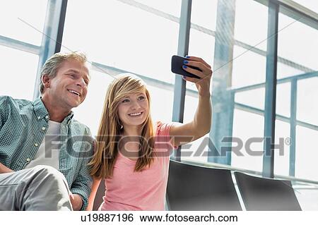 Stock Photograph - Portrait of young beautiful teenage girl taking selfie with her father in airport. Fotosearch