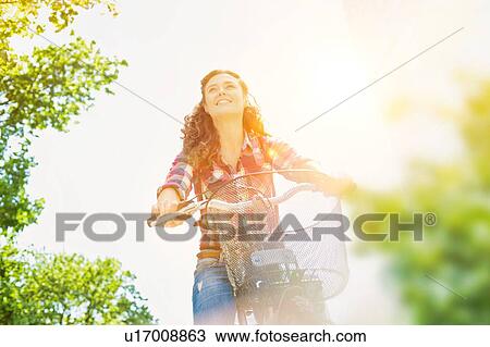 Stock Image - Portrait of young beautiful woman smiling while riding bicycle in the middle of the city with lens flare in background. Fotosearch