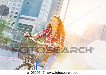 Stock Photo - Portrait of young beautiful woman smiling while riding bicycle in the middle of the city with lens flare in background. Fotosearch