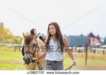 Stock Photo - Portrait of young beautiful woman standing with horse in ranch. Fotosearch