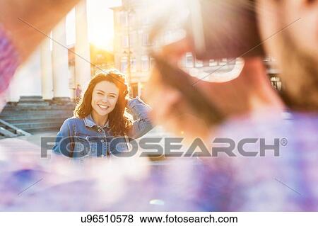 Stock Photo - Portrait of young beautiful woman posing while being photograph by his boyfriend with lens flare in background. Fotosearch