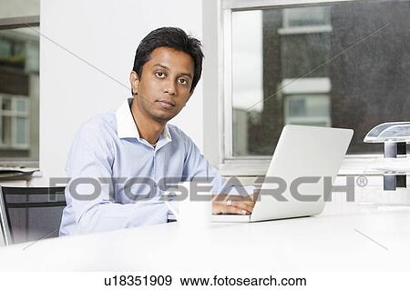 Stock Photo - Portrait of young businessman using laptop at desk in office. Fotosearch