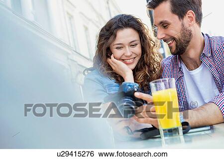 Stock Photograph - Portrait of young handsome man showing pictures on camera to his girlfriend while waiting for their food . Fotosearch