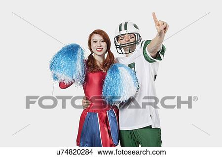 Picture - Portrait of young man in football uniform and cheerleader with pom-pom standing over gray background. Fotosearch