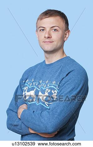 Stock Photo - Portrait of young man in sweater with arms crossed against blue background. Fotosearch