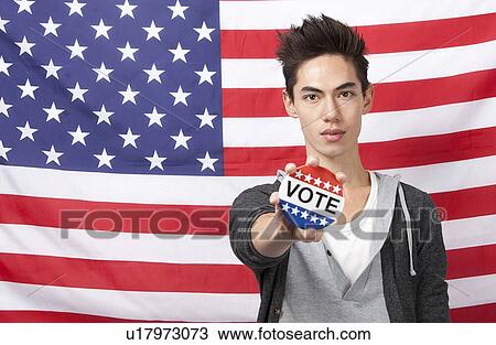 Stock Image - Portrait of young man showing vote badge standing against American flag. Fotosearch