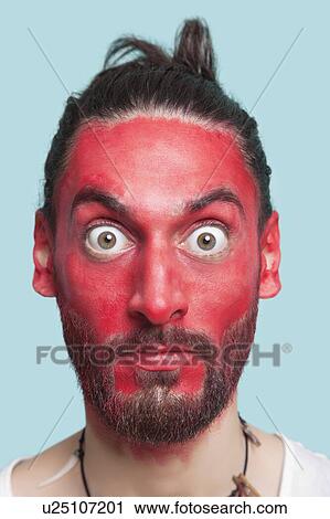 Portrait of young man with red painted face and wide eyes against blue background View Large Photo Image Stock Image - Portrait of young man with red painted face and wide eyes against blue background. Fotosearch