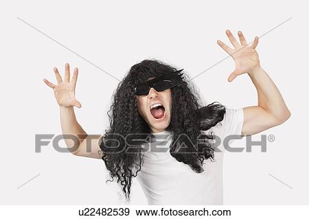 Stock Photo - Portrait of young man with wig and sunglasses shouting against gray background. Fotosearch