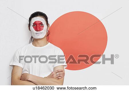 Stock Image - Portrait of young mixed race person with painted face and arms crossed against Japanese flag. Fotosearch