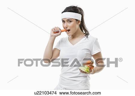 Picture - Portrait of young sportswoman eating carrot over white background. Fotosearch
