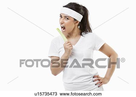 Stock Image - Portrait of young sportswoman eating celery over white background. Fotosearch