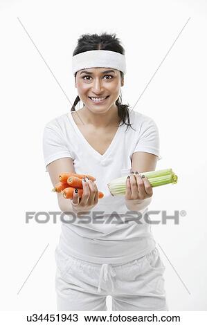 Stock Image - Portrait of young sportswoman holding carrots and celery over white background. Fotosearch