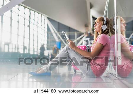 Stock Image - Portrait of young teenage girl sitting on the floor while watching movie on her digital tablet with headphones on in airport. Fotosearch