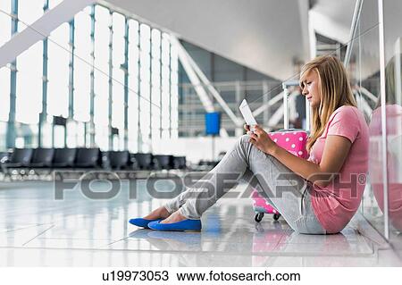 Stock Image - Portrait of young teenage girl sitting on the floor and using digital tablet while waiting for her flight in airport. Fotosearch