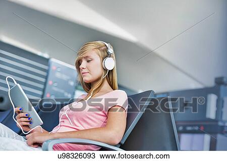 Stock Image - Portrait of young teenage girl sitting while watching movie on her digital tablet with headphones on in airport. Fotosearch