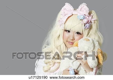 Stock Image - Portrait of young woman dressed as a doll holding soft toy over gray background. Fotosearch