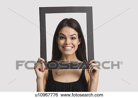 Stock Photograph - Portrait of young woman smiling through frame over white background. Fotosearch