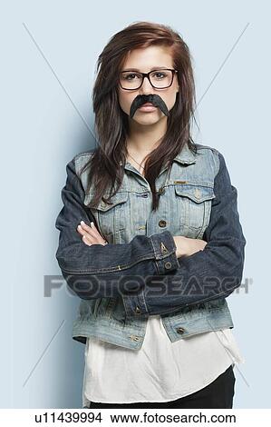 Picture - Portrait of young woman wearing fake mustache with arms crossed against light blue background. Fotosearch