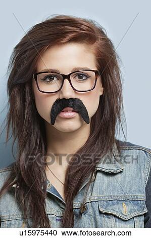 Stock Image - Portrait of young woman wearing eyeglasses and fake mustache against light blue background. Fotosearch