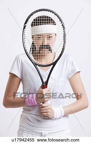 Stock Photography - Portrait of young woman with a fake mustache holding tennis racket in front of her face against white background. Fotosearch
