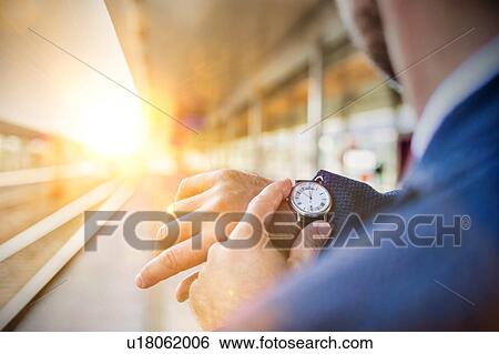 Stock Photograph - Rear view of businessman looking at his watch while waiting in train station. Fotosearch