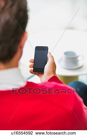 Stock Image - Rear view of businessman using smartphone while sitting and drinking coffee in lobby. Fotosearch