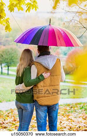 Stock Image - Rear view of man holding umbrella while enjoying the lovely view with his wife in park. Fotosearch