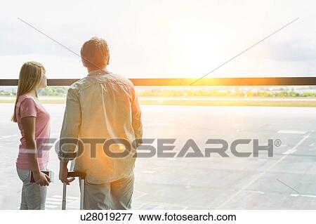Stock Photo - Rear view of mature man looking through the window with his daughter in airport. Fotosearch
