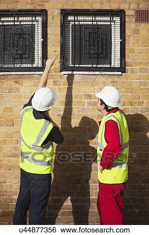 Stock Photograph - Rear view of two male worker in hardhats pointing towards windows. Fotosearch