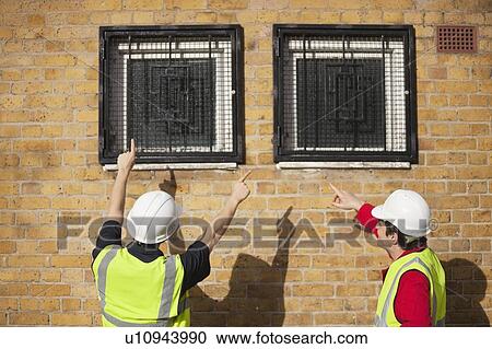 Rear view of two male workers in hardhats pointing towards windows View Large Photo Image Stock Image - Rear view of two male workers in hardhats pointing towards windows. Fotosearch