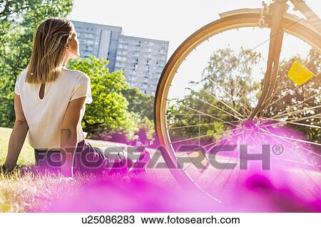 Stock Image - Rear view of young attractive businesswoman sitting on the grass while watching the sunset. Fotosearch