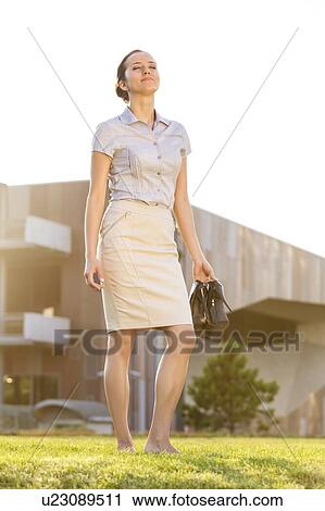 Stock Image - Relaxed young businesswoman holding footwear in office lawn. Fotosearch