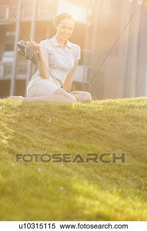 Stock Photography - Relaxed young businesswoman holding high heels while sitting in office lawn. Fotosearch