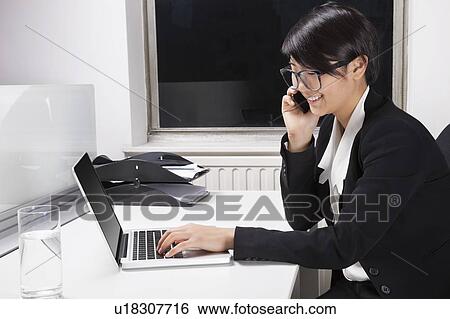 Side view of young businesswoman using laptop and cell phone at desk in office View Large Photo Image Stock Photograph - Side view of young businesswoman using laptop and cell phone at desk in office. Fotosearch