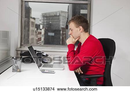 Picture - Side view of young man using laptop at desk in office. Fotosearch